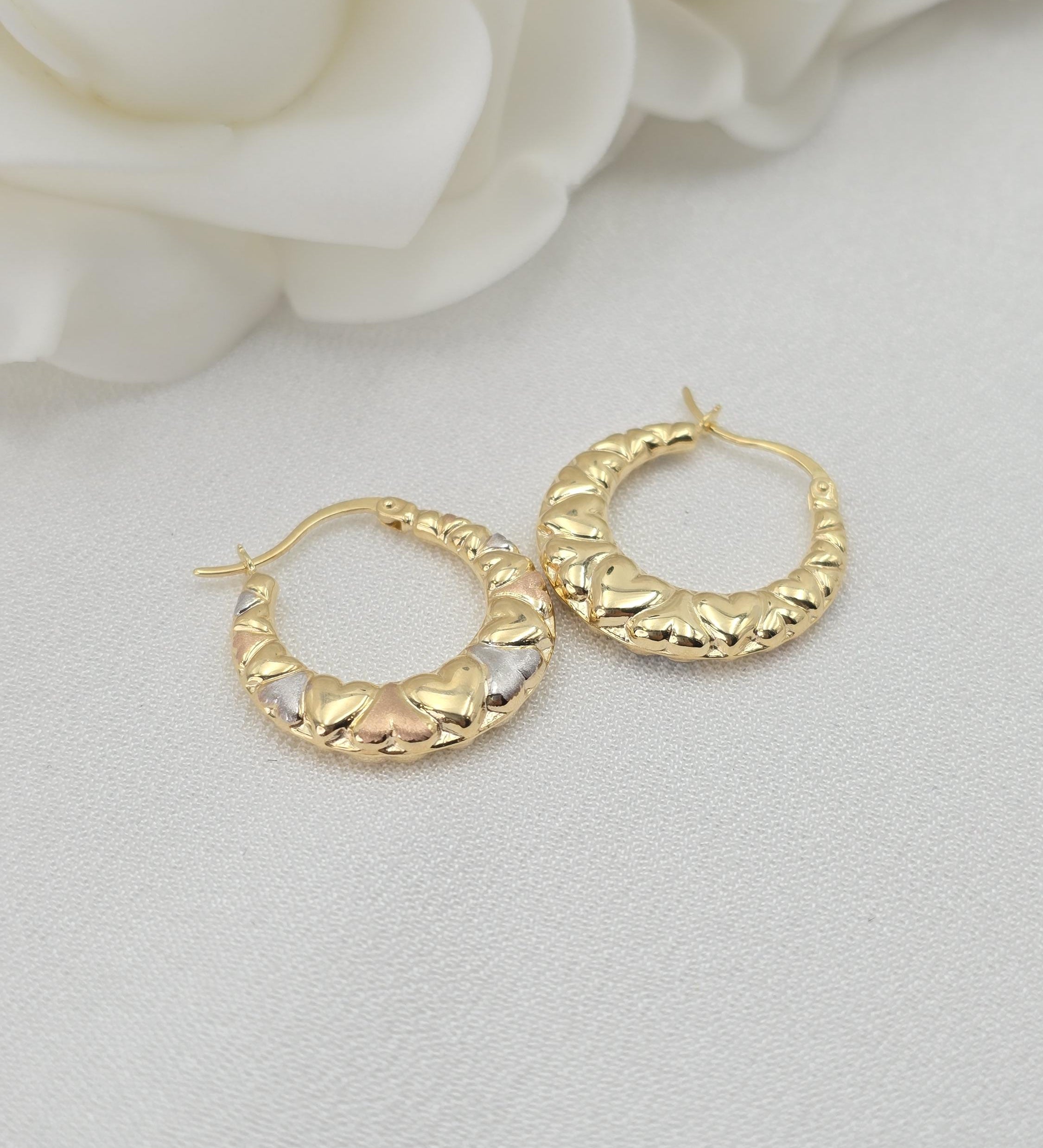 May include: Close-up of a pair of gold hoop earrings. The hoops are adorned with small, heart-shaped details in three-toned gold: yellow, white, and rose. The earrings have a smooth, curved design and are displayed against a white background.