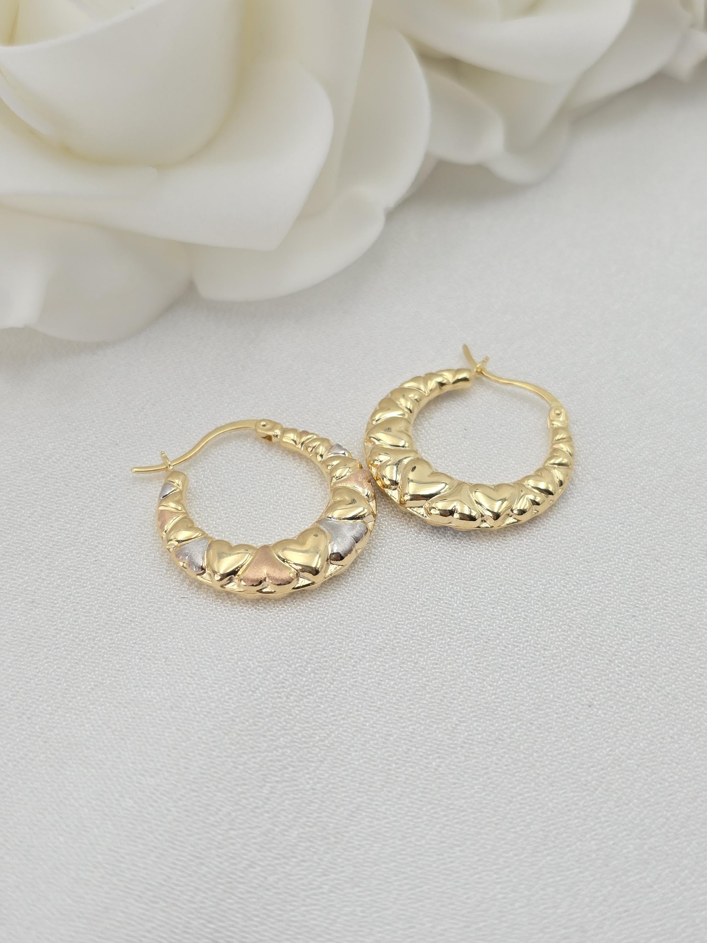 May include: Close-up of a pair of gold hoop earrings. The hoops are adorned with small, heart-shaped details in three-toned gold: yellow, white, and rose. The earrings have a smooth, curved design and are displayed against a white background.